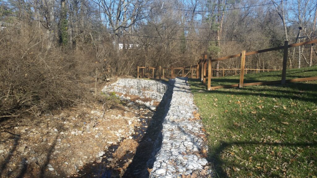 Stone-lined drainage channel beside a wooden fence in a wooded residential area.