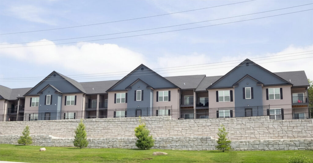 Modern gray apartment buildings behind a tiered stone retaining wall and landscaped lawn.