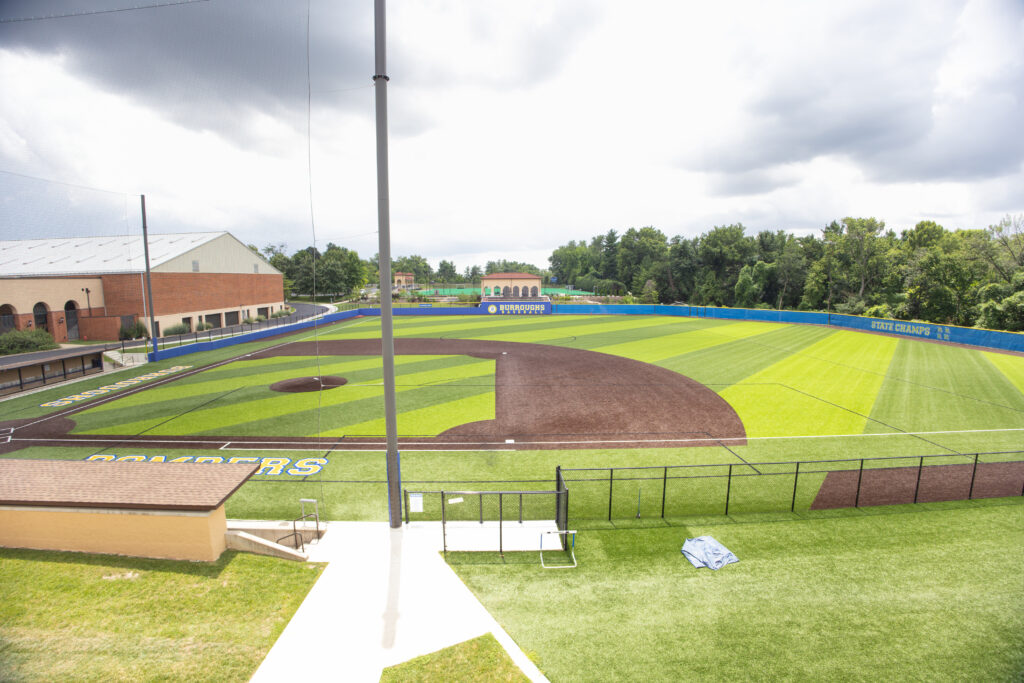 Burroughs School baseball field with striped artificial turf, blue outfield walls, and an adjacent athletic facility.